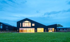Contemporary black timber barn-style house with floor-to-ceiling glass windows glowing at dusk, featured in Architecture Vogue magazine