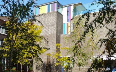 Contemporary brick residential building with colorful glass facade panels and rooftop terrace, surrounded by trees in an urban courtyard